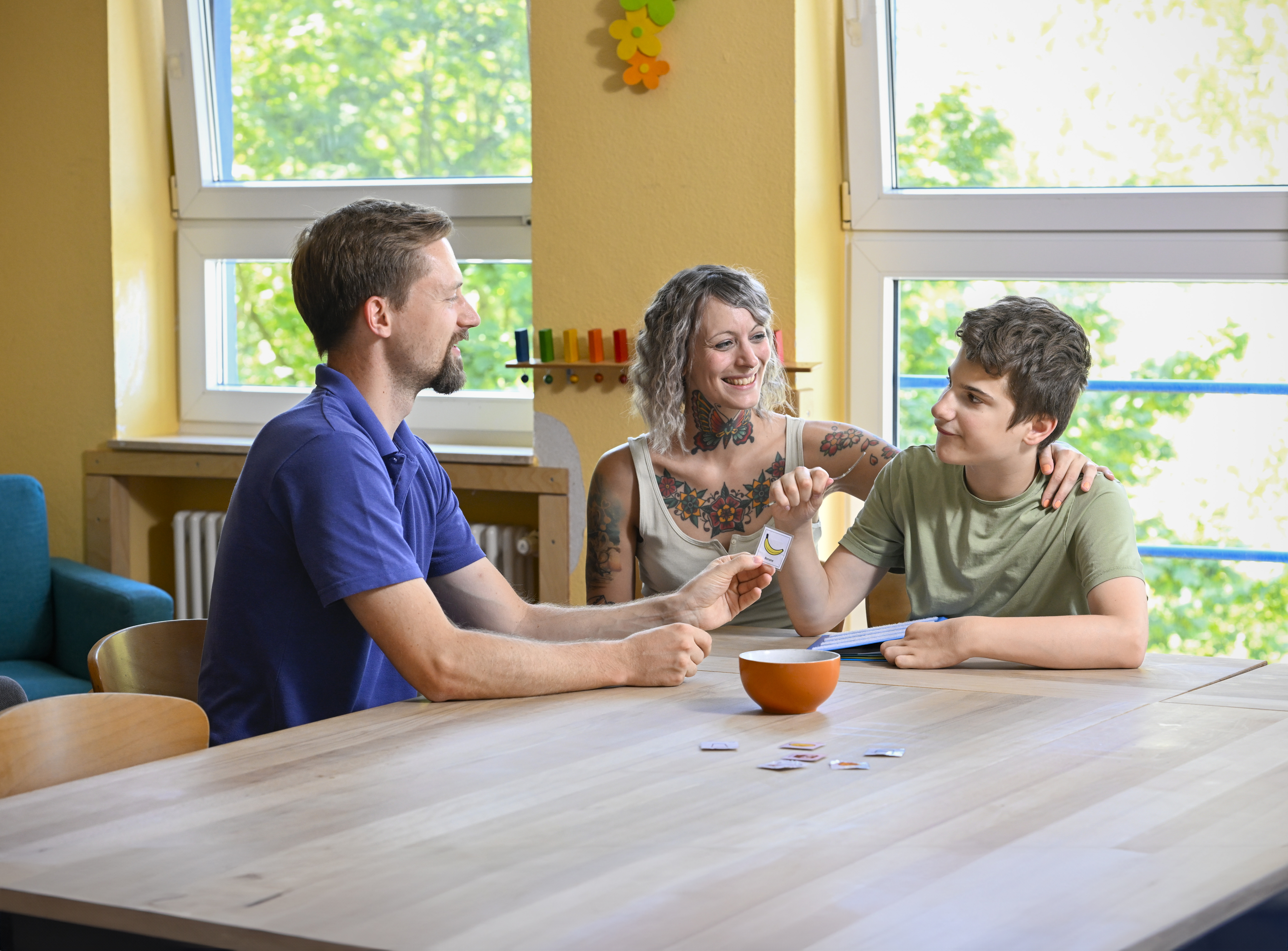 Drei Personen sitzen an einem Tisch in einem hellen Raum und machen gemeinsam eine Übung mit Bildkarten.