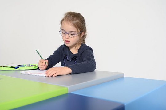 Kind mit Brille schreibt an einem Tisch in einem modernen Raum.