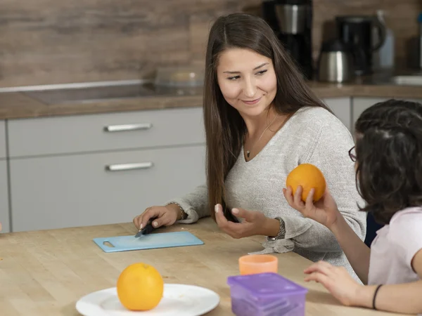 Frau sitzt mit Kindern an einem Tisch; sie schälen eine Orange und unterhalten sich.