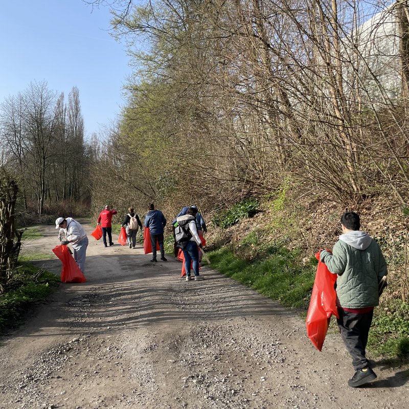 Auf einem Weg sind mehrere Menschen zu sehen, die rote Müllsäcke und Greifzangen haben und Müll einsammeln.