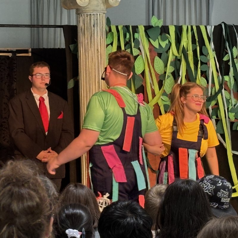 Szenenbild: Papageno und Papagena tanzen mit eingehakten Armen. Links auf der Bühne steht Fürst Sarastro.