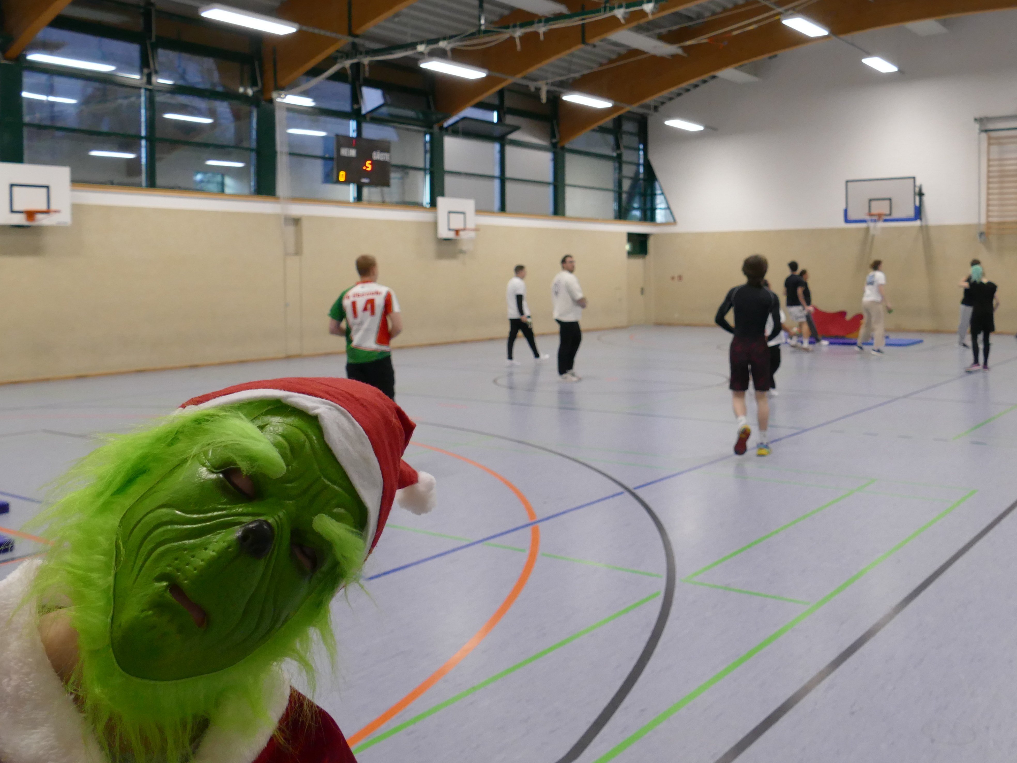 In einer Sporthalle bewegen sich im Hintergrund Schüler. Vorne hält eine Person mit einer grünen "Grinch"-Maske den Kopf ins Bild