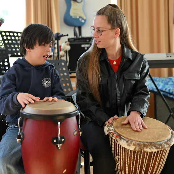 Ein Junge und eine junge Frau sitzen im Musikraum der Schule und haben große Trommeln vor sich. Die Frau sieht zu dem Jungen hin.