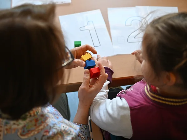 Die I-Helferin zeigt einem Mädchen drei bunte Bausteine. Die Kamera schaut von hinten oben auf die beiden. Vor ihnen auf dem Tisch liegen Blätter mit den Zahlen 1 und 2.