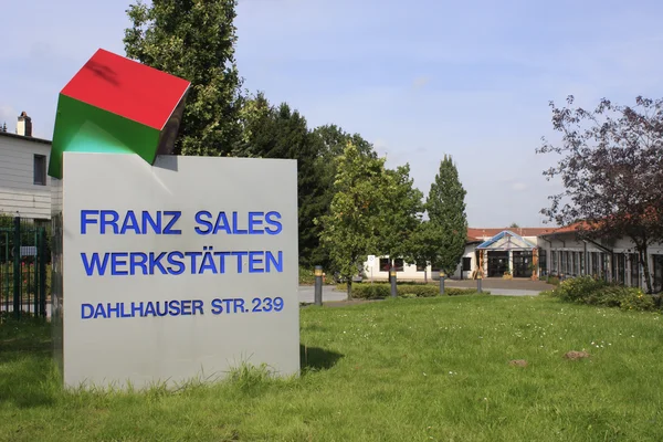 Schild mit "Franz Sales Werkstätten", grüne Wiese und Gebäude im Hintergrund.