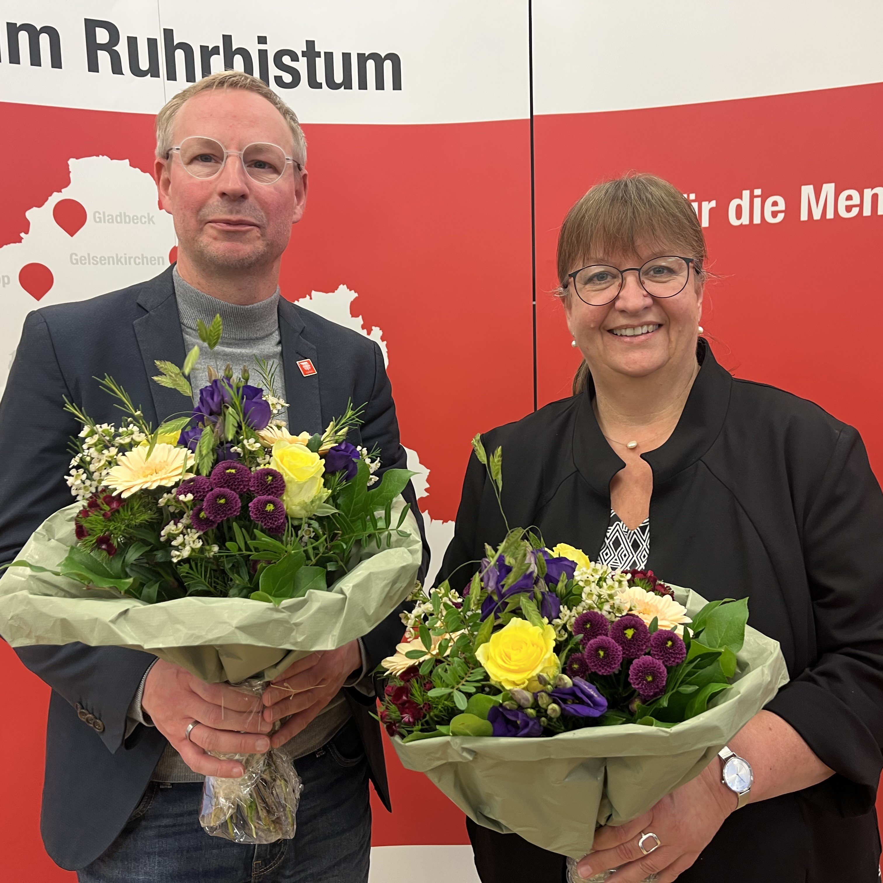 Die neu gewählten Ratsmitglieder Michael Kreuzfelder und Nicole Köster stehen vor einer Rückwand mit Caritas-Beschriftung und haben Blumensträuße in den Händen
