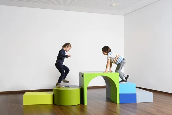Kinder spielen auf bunten Kletterformen in einem leeren Raum.