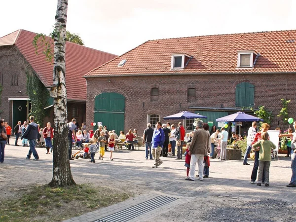 Menschenmenge bei einer Veranstaltung auf einem Bauernhof mit roten Backsteingebäuden.