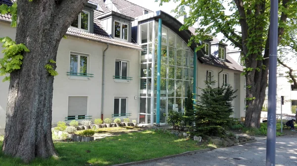 Modernes Gebäude mit Glasfassade, Bäumen und kleinem Garten im Vordergrund.