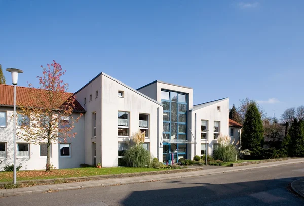 Modernes Gebäude mit Glasfassade, blauer Himmel, Bäume und Straße im Vordergrund.