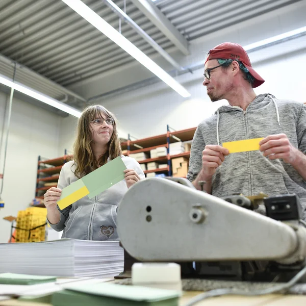 Zwei Personen in einer Werkstatt halten bunte Papierstreifen in den Händen