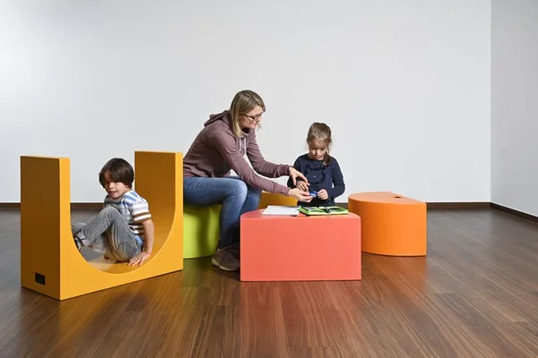 Erwachsene und zwei Kinder mit bunten Sitzwürfeln in leerem Raum.