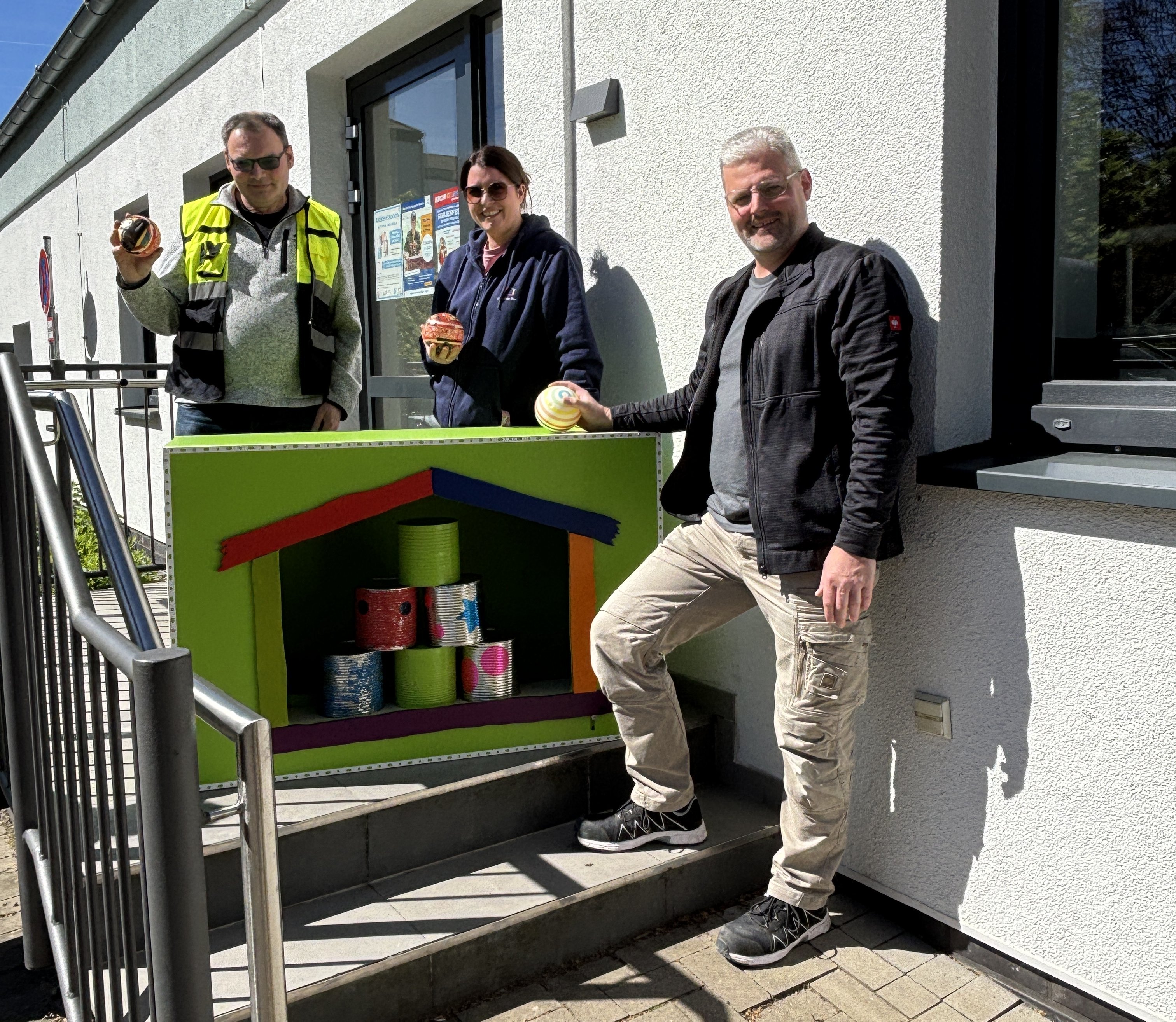 Zwei Männer und eine Frau präsentieren eine große Kiste aus Holz, bei der vorne eine Haus-Form ausgeschnitten ist. Darin stehen Dosen zum Dosenwerfen. Die Personen halten die Bälle zum Dosenwerfen in der Hand.