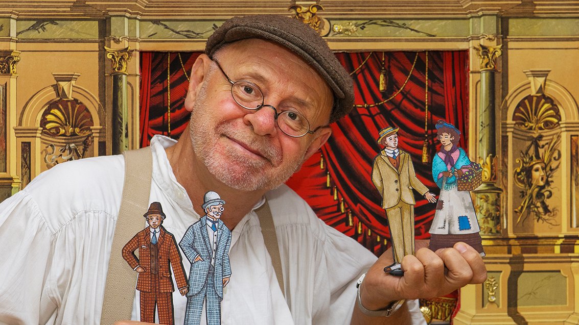 Mann in Mütze mit Papiertheaterfiguren vor dekorativer historischer Bühne.