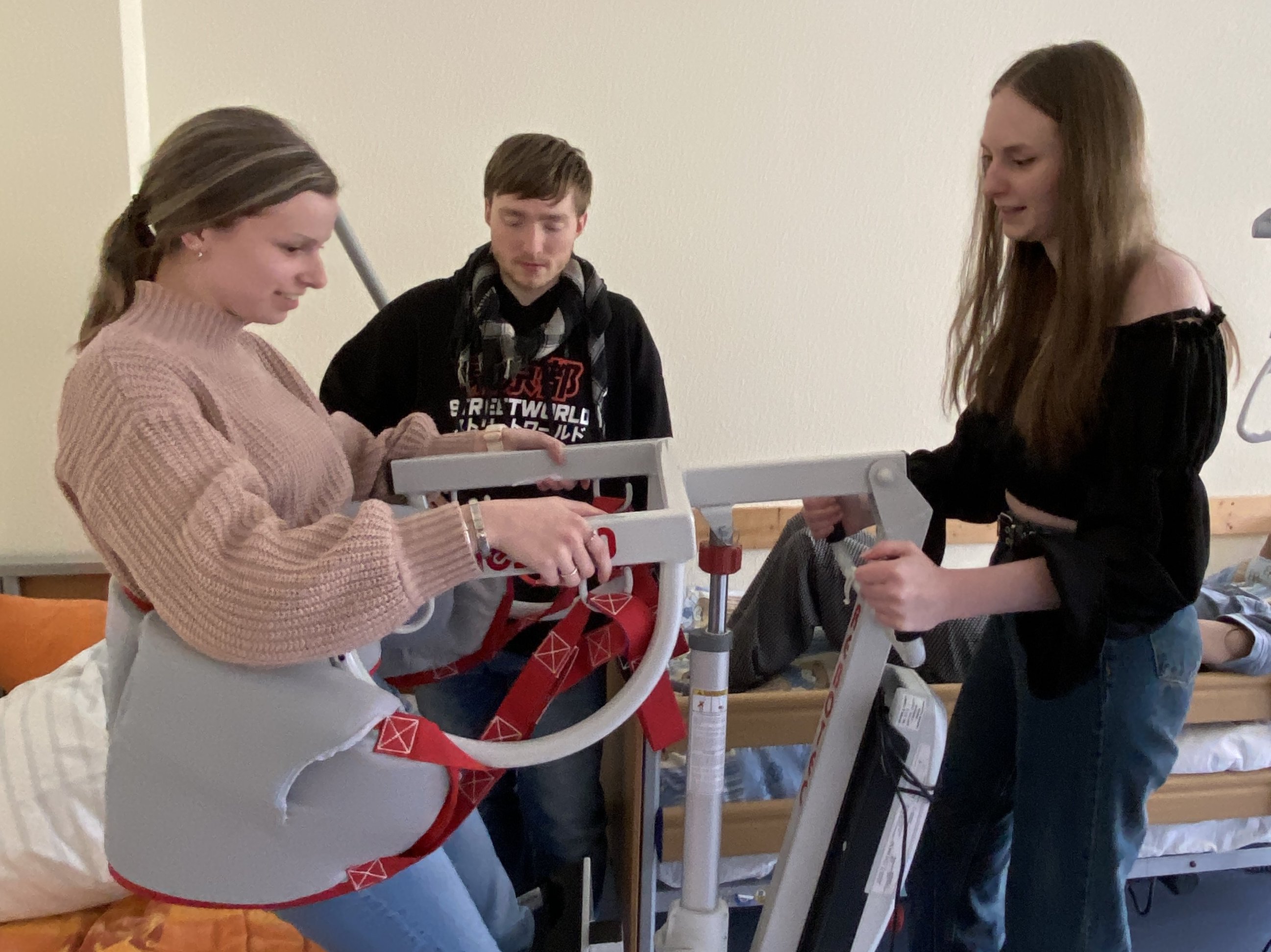 Eine junge Frau hilft einer anderen jungen Frau bei der Benutzung eines medizinischen Lifters in einem Zimmer. Ein junger Mann beobachtet sie in der Nähe.