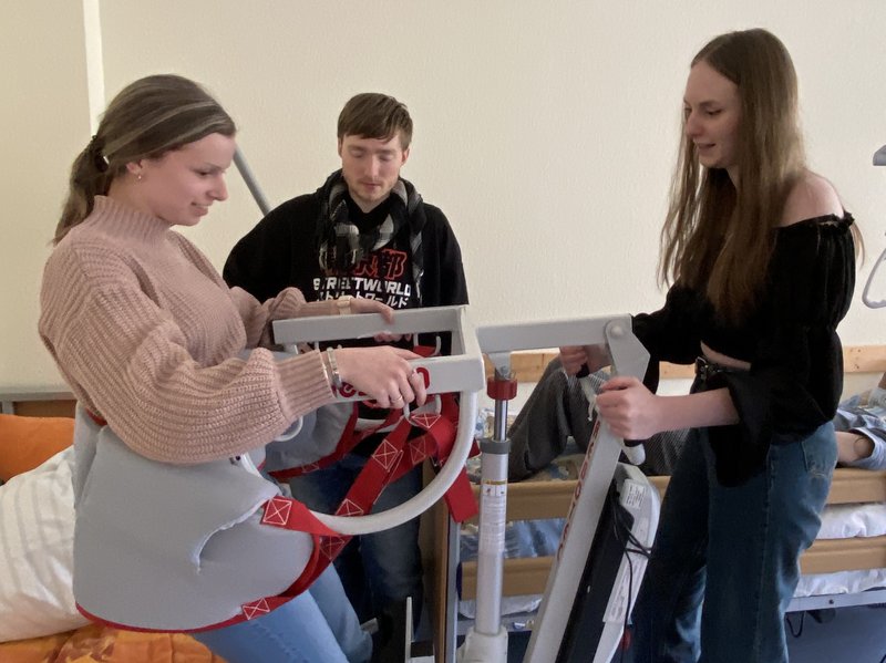 Eine junge Frau hilft einer anderen jungen Frau bei der Benutzung eines medizinischen Lifters in einem Zimmer. Ein junger Mann beobachtet sie in der Nähe.