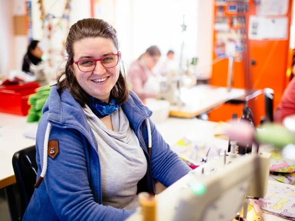 Frau lächelt in einer Werkstatt, Nähmaschinen im Hintergrund.