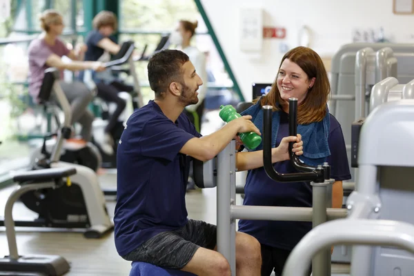 Menschen trainieren an Fitnessgeräten in einem Fitnessstudio, Mann und Frau lachen dabei.