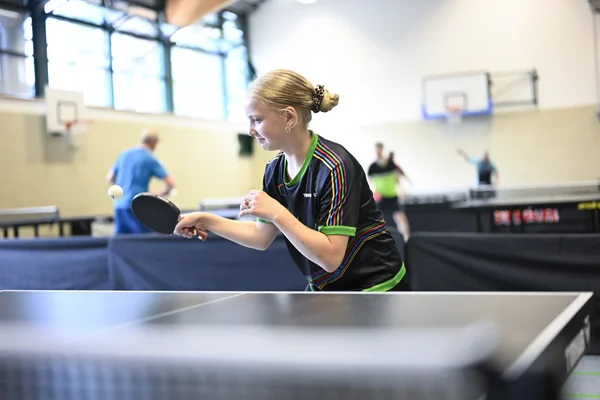 Mädchen spielt Tischtennis in einer Sporthalle, verschwommener Hintergrund mit anderen Spielern.