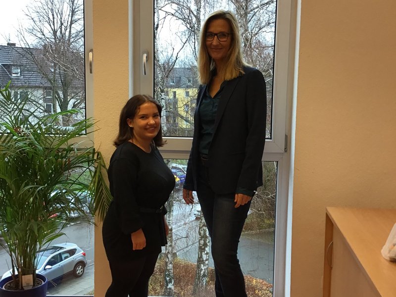 Zwei Personen stehen vor einem Fenster, neben einer Pflanze im Innenraum.