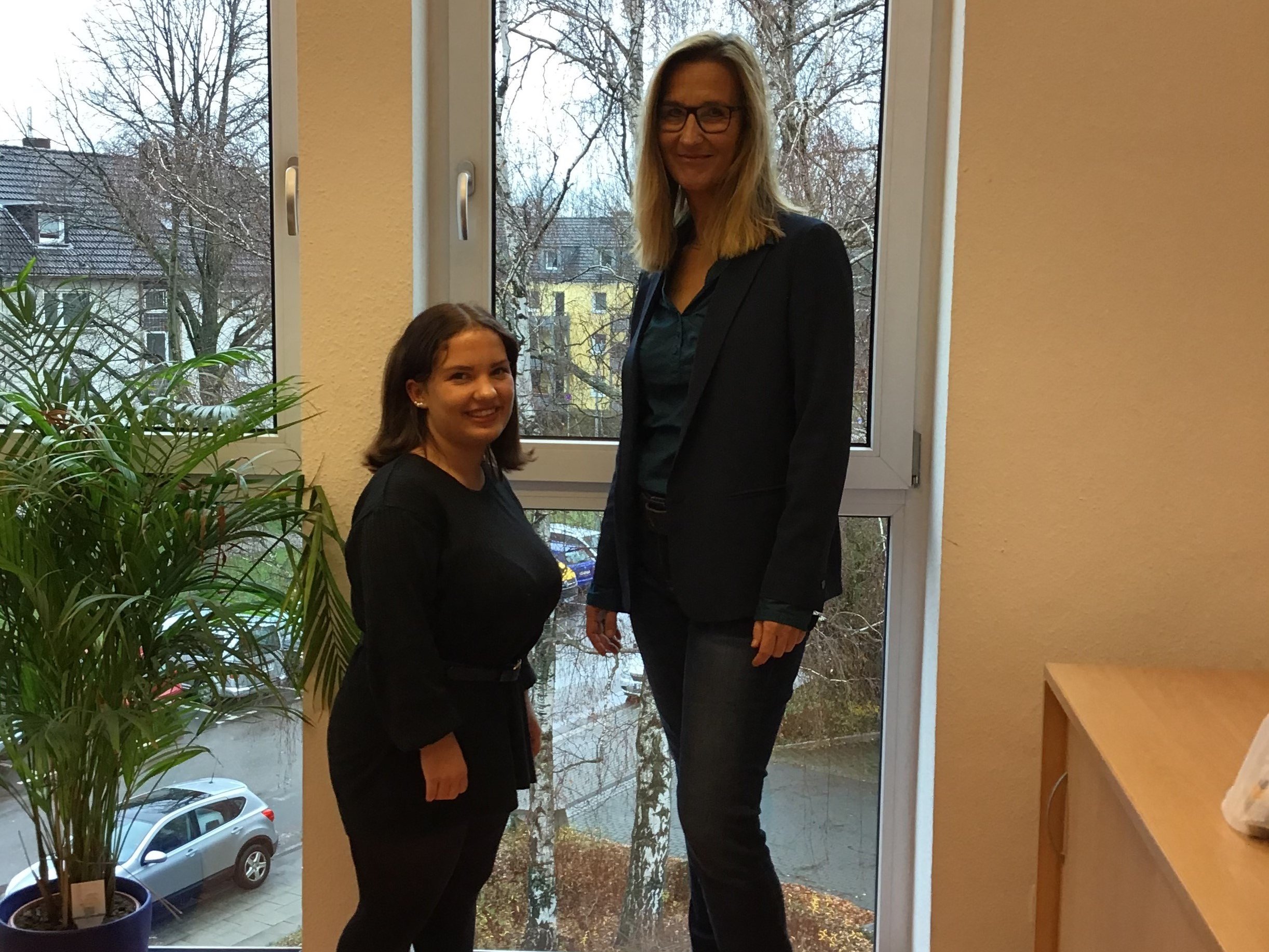 Zwei Personen stehen vor einem Fenster, neben einer Pflanze im Innenraum.