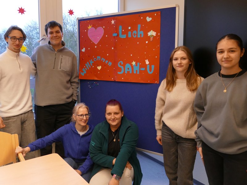 Schüler aus dem Oberkurs stehen vor einem Plakat mit der Aufschrift "Herzlich willkommen SAH-U". Damit ist der neue Unterkurs der Sozialassistenten gemeint.