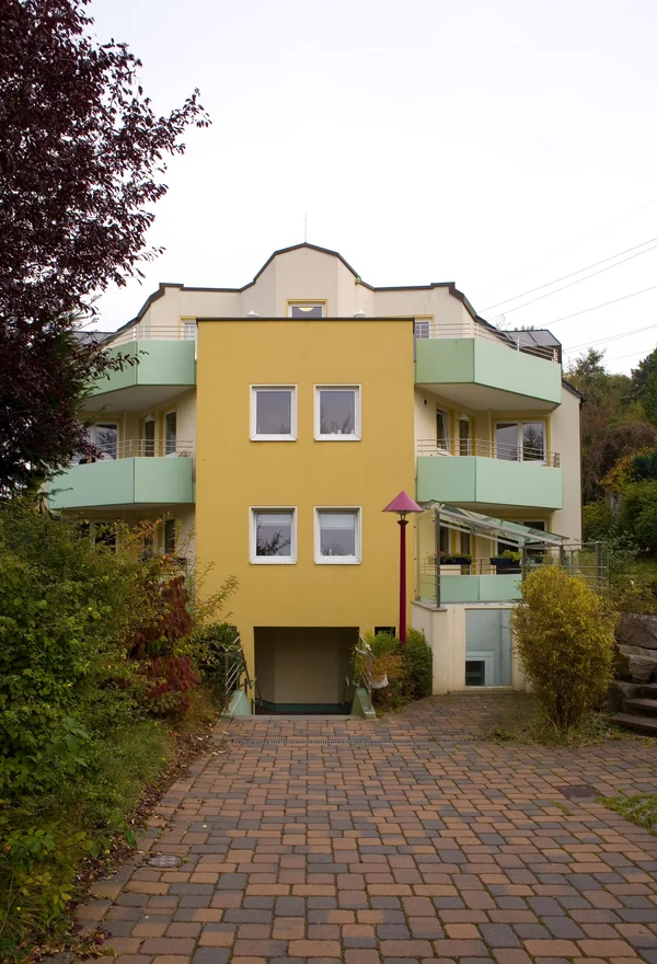 Gelbes Gebäude mit Balkonen, umgeben von Bäumen und einer gepflasterten Einfahrt.