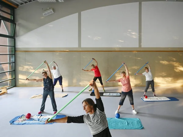 Menschen im Fitnesskurs strecken sich mit Stöcken in einem hellen Raum.
