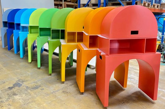 Bunte Sitzbänke in Regenbogenfarben, aufgereiht in einer Lagerhalle.