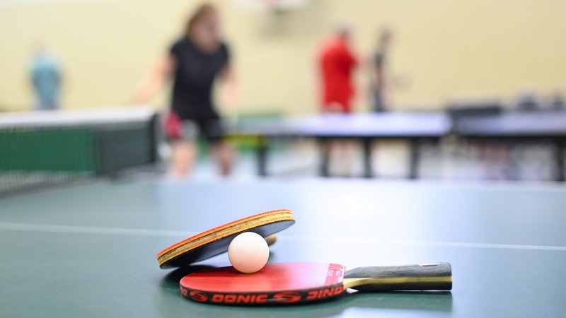 Zwei Tischtennisschläger und ein Ball liegen auf einer Tischtennisplatte. Im Hintergrund sind unscharf weitere Tischtennisplatten zu sehen, an denen gespielt wird.