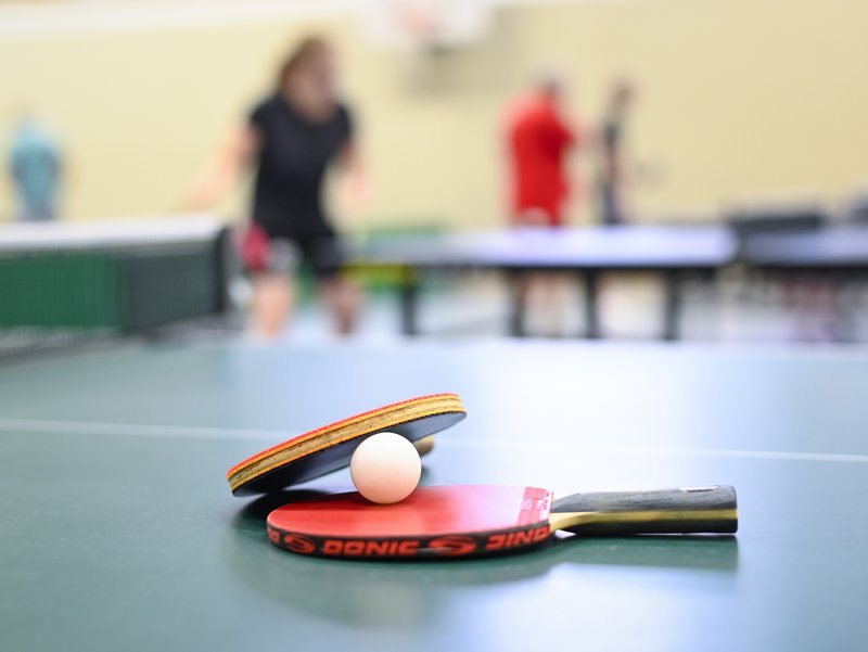 Tischtennisschläger und Ball liegen auf dem Tisch. Im Hintergrund sieht man unscharf Tischtennisspieler beim Training