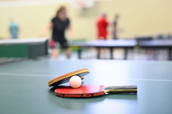 Zwei Tischtennisschläger und ein Ball liegen auf einer Tischtennisplatte. Im Hintergrund sind Tischtennisspieler an weiteren Platten zu sehen.