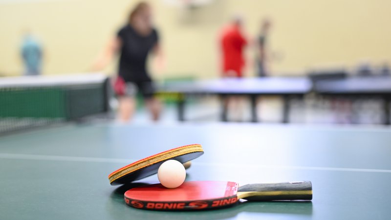 Tischtennisschläger und Ball liegen auf dem Tisch. Im Hintergrund sieht man unscharf Tischtennisspieler beim Training