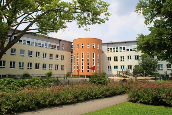 Gebäude mit rundem, orangenem Treppenturm, umgeben von Grünflächen und Bäumen.