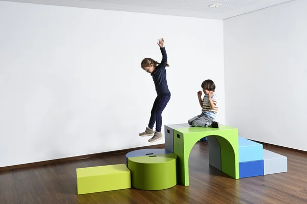 Kinder spielen auf bunten geometrischen Spielblöcken in einem leeren Raum.