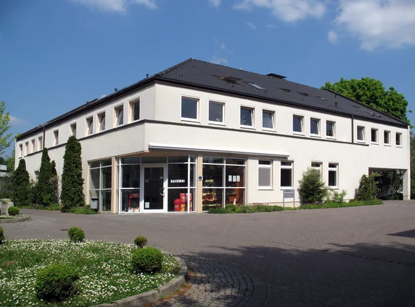 Modernes zweistöckiges Gebäude mit Glasfront, umgeben von Bäumen und Rasenflächen.