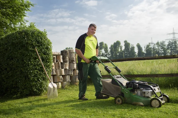 Mann mäht Rasen mit Benzinrasenmäher, Schaufel lehnt an Hecke.