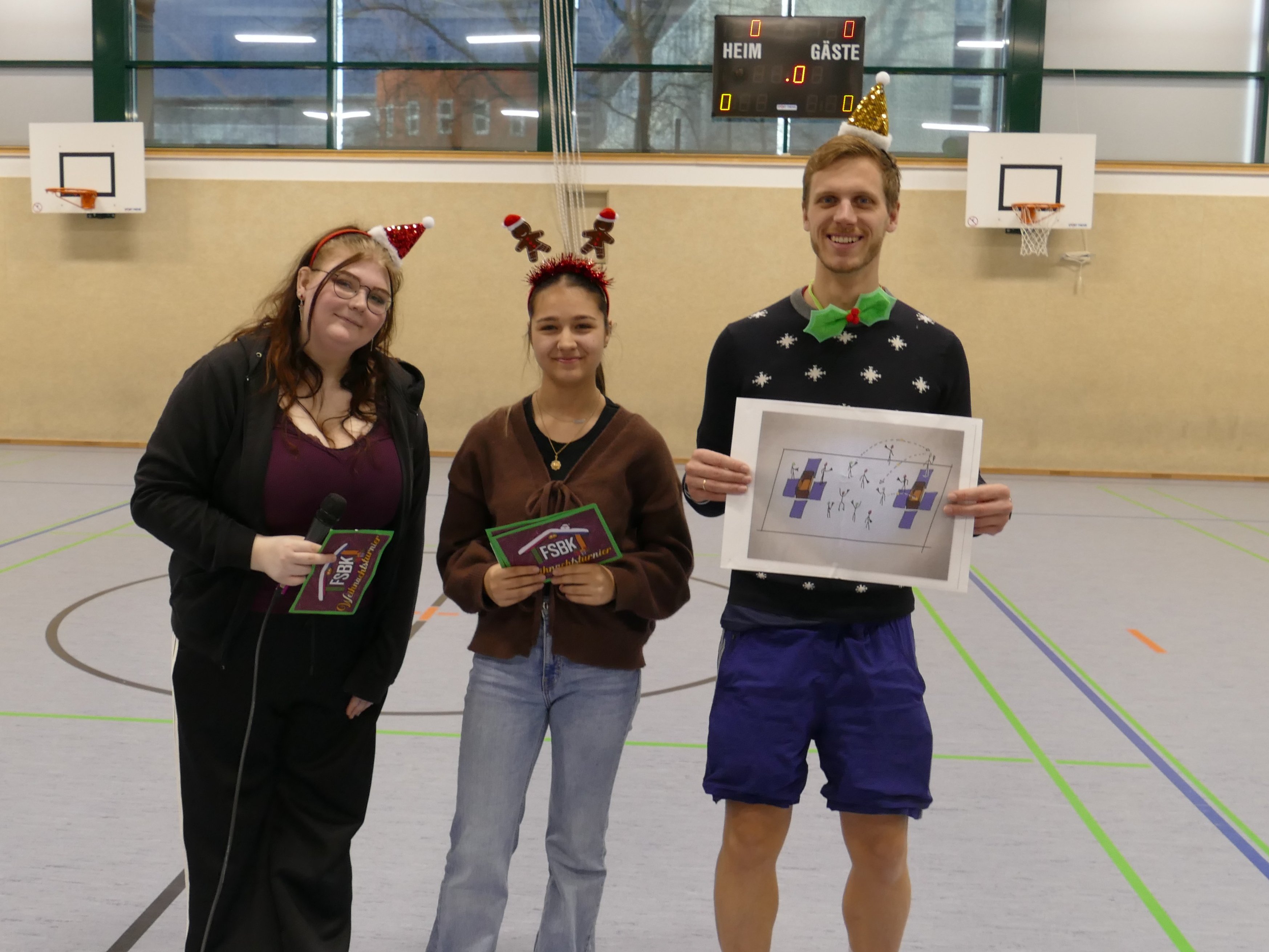 Lehrer Nils Eißing und zwei Schülerinnen haben weihnachtliche Haar-Reifen auf dem Kopf und blicken in die Kamera. Sie halten Unterlagen zu dem sportlichen Spiel in der Hand, das sie sich für das Weihnachtsturnier ausgedacht haben.