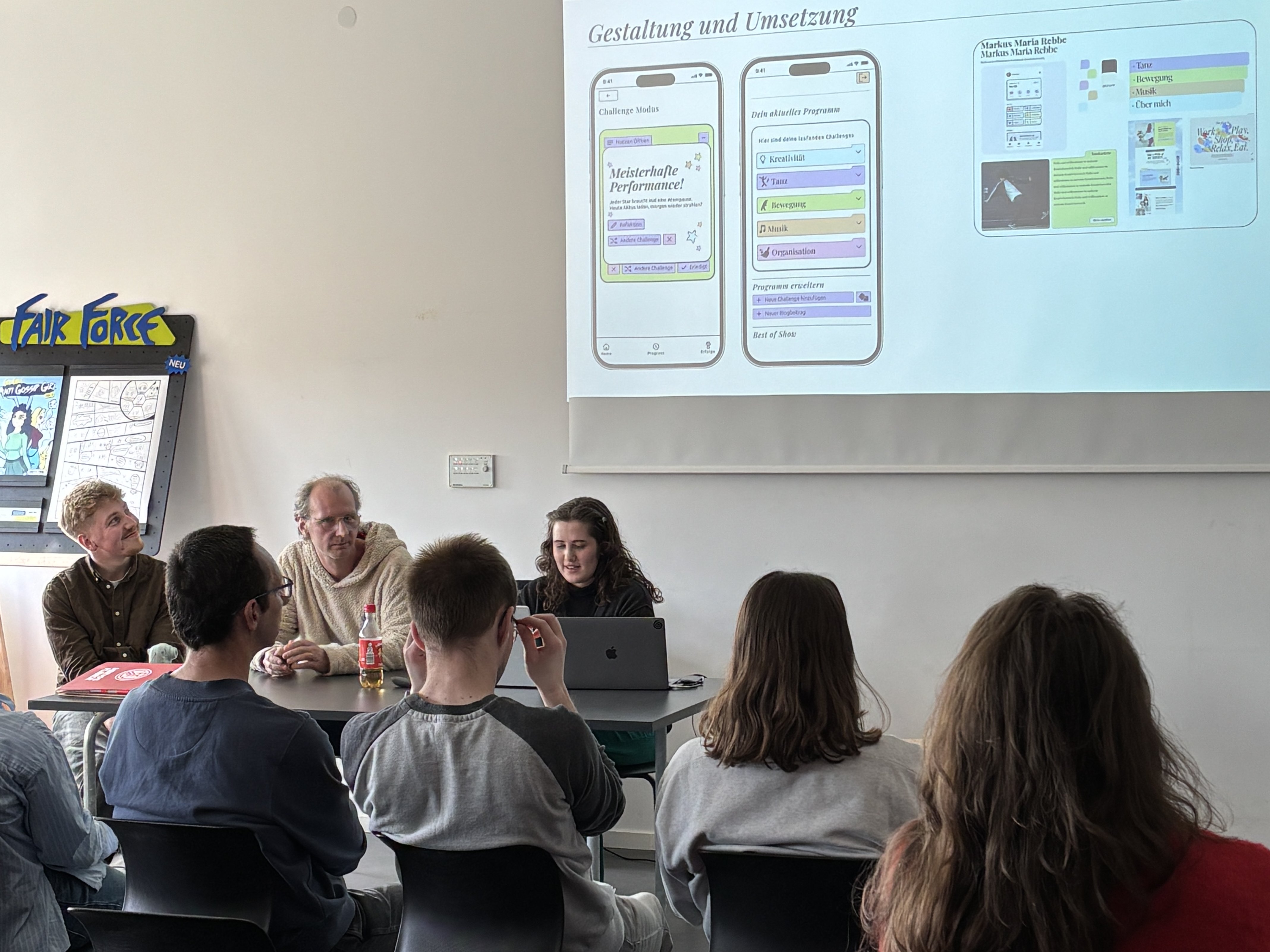 Co-Designer Markus und Studentin Katja bei ihrer Präsentation. Auf der Leinwand an der Wand ist die entwickelte App dargestellt.