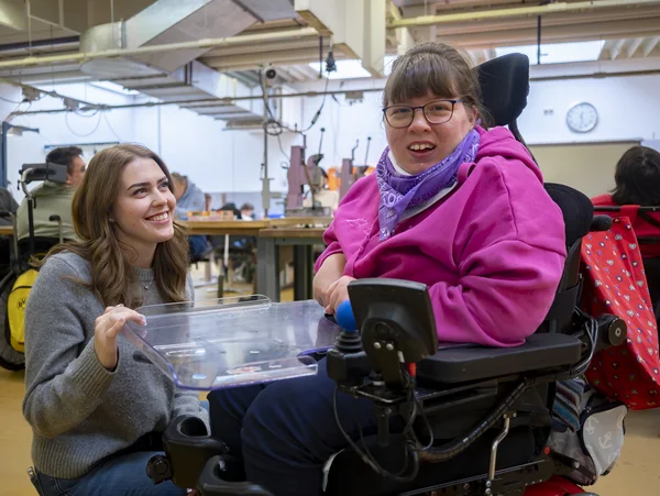 Frau im Rollstuhl und kniende Frau in Werkstatt, beide lächeln.