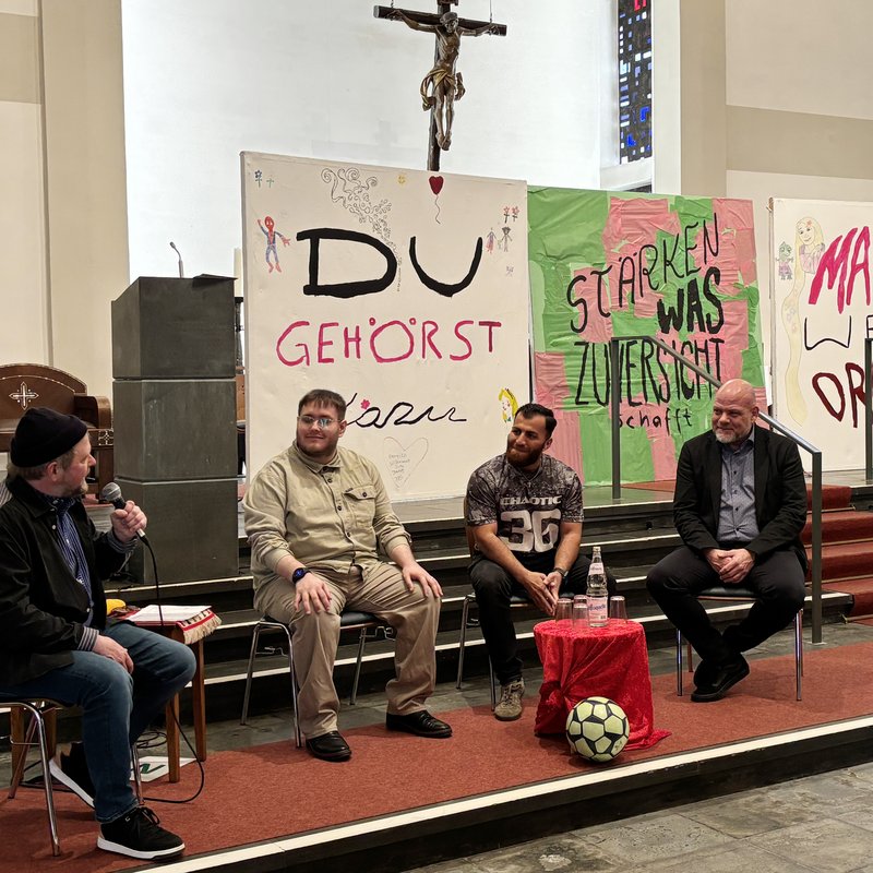 Gesprächsrunde in der Kirche des Franz Sales Hauses. Der Moderator und drei Diskussionsteilnehmer sitzen auf Stühlen. Im Hintergrund stehen große Bilder, auf denen das Motto des Josefstags geschrieben steht.