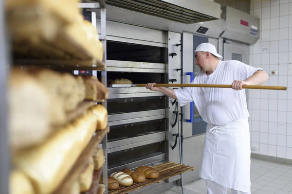 Bäcker schiebt Brot mit einem Schieber in Ofen; Regale voller Brot im Vordergrund.
