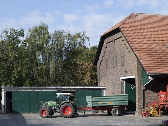 Traktor mit Anhänger vor Scheune und Bäumen bei bewölktem Himmel.