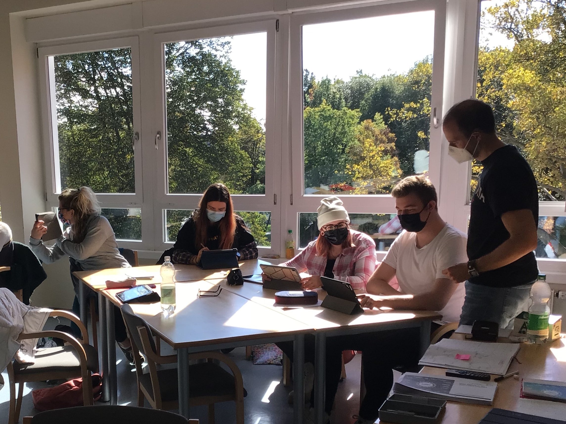 Menschen arbeiten in einem sonnigen Raum an Tablets und Laptops, Bäume sind draußen zu sehen.