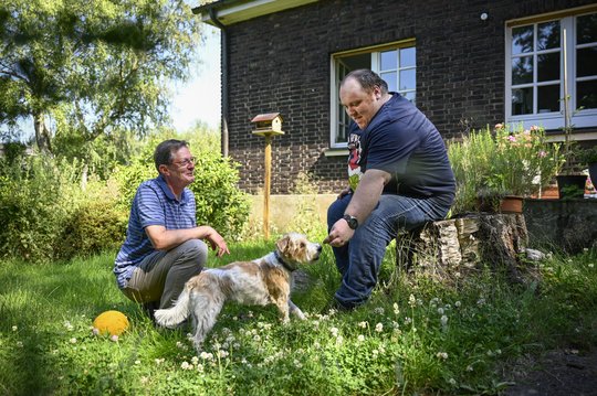 Zwei Männer und ein Hund im Garten vor einem schönen Wohnhaus.