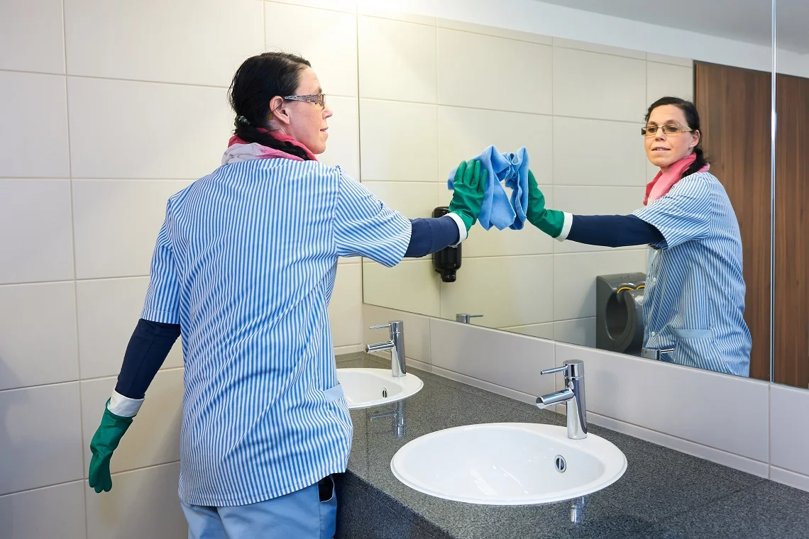 Mitarbeiterin der Gebäudereinigung säubert einen Spiegel in einem Sanitärbereich – stellvertretend für Reinigungsdienste.